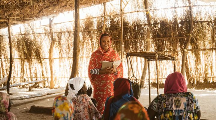 A woman sharing the gospel with a group seated in a refugee camp shelter