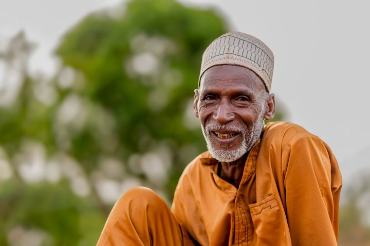 A smiling elderly man in orange robes and a white cap